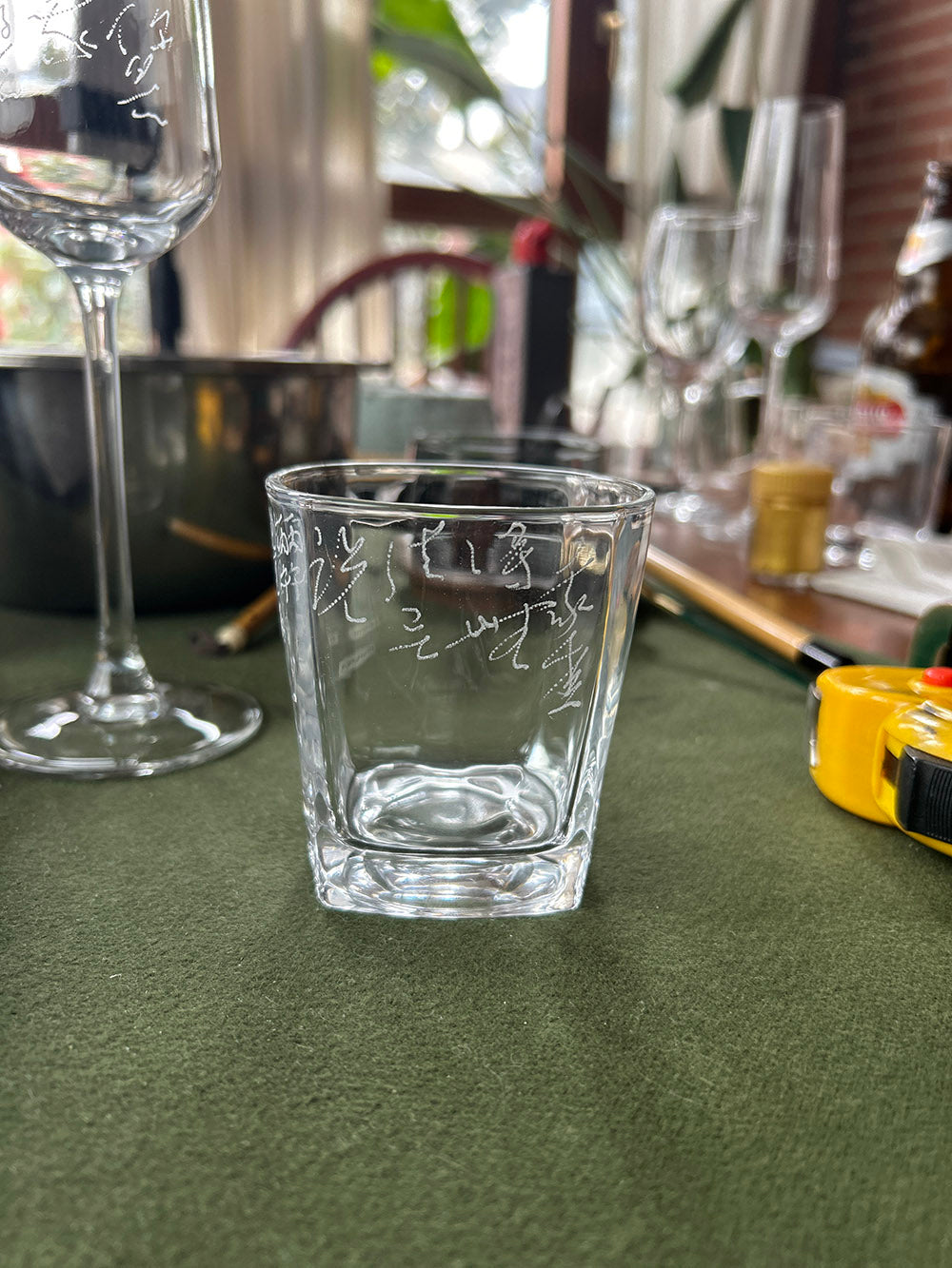 Full shot of square hand‑engraved calligraphy tumbler glass placed on green fabric table surface