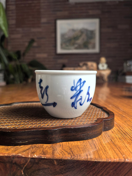 Hand-thrown Jingdezhen qingbai porcelain cylindrical tea cup with "Joy" calligraphy, placed on a woven bamboo mat in a traditional zen tea setting with wooden background