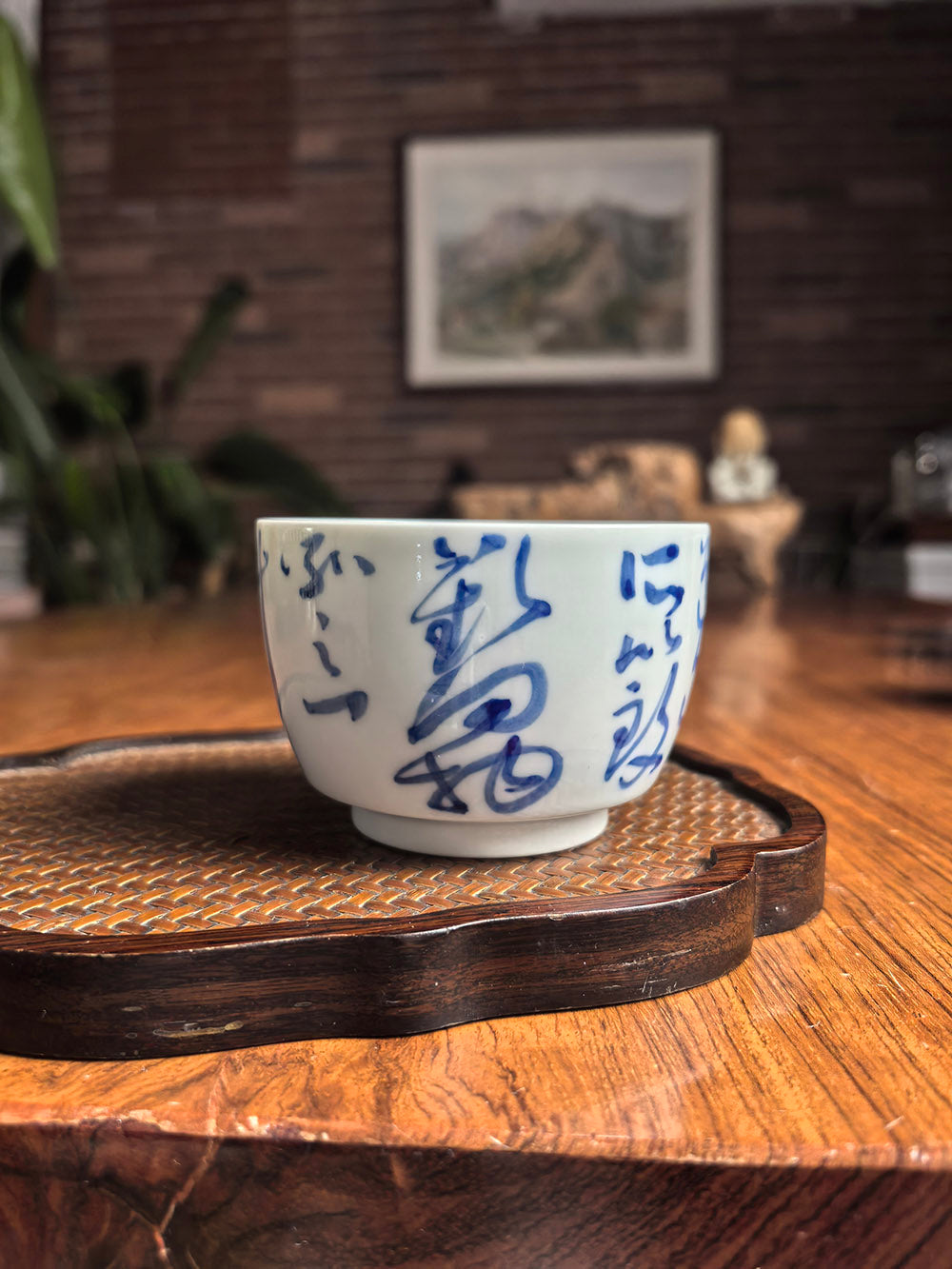 Hand-thrown Jingdezhen qingbai porcelain cylindrical tea cup with "Joy" calligraphy, placed on a woven bamboo mat in a traditional zen tea setting with wooden background