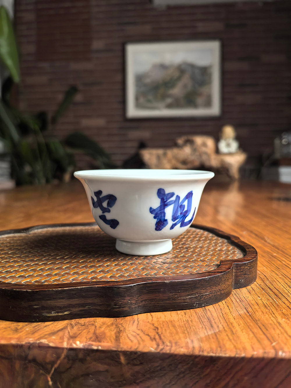 Hand-thrown Jingdezhen qingbai porcelain "press-hand" tea cup with "watching clouds listening to rain" calligraphy, placed on woven bamboo mat in zen tea setting with landscape painting and wooden statue