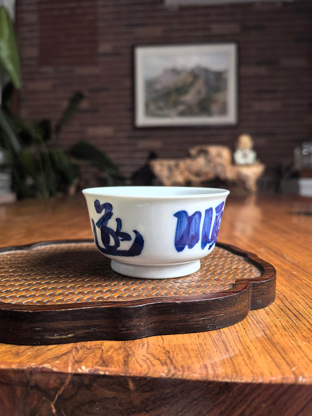Hand-thrown Jingdezhen qingbai porcelain flat tea cup with "Smooth Sailing" calligraphy, placed on a woven bamboo mat in a zen tea setting with landscape painting background