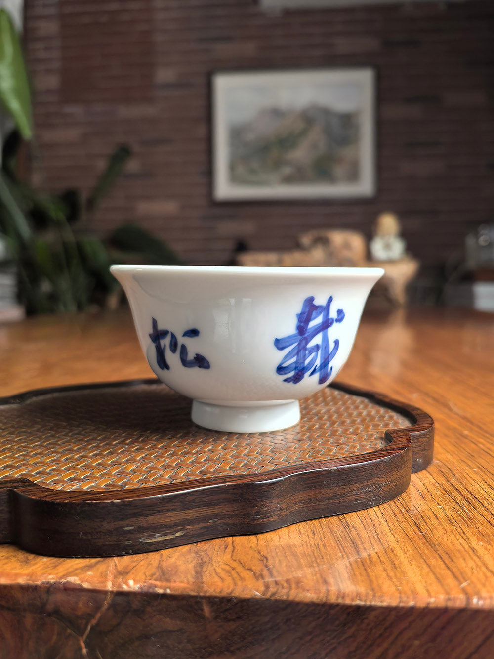 Hand-thrown Jingdezhen qingbai porcelain press-hand tea cup with "Planting Pines for Great Harmony" calligraphy, placed on a dark wood tea table with bamboo mat background