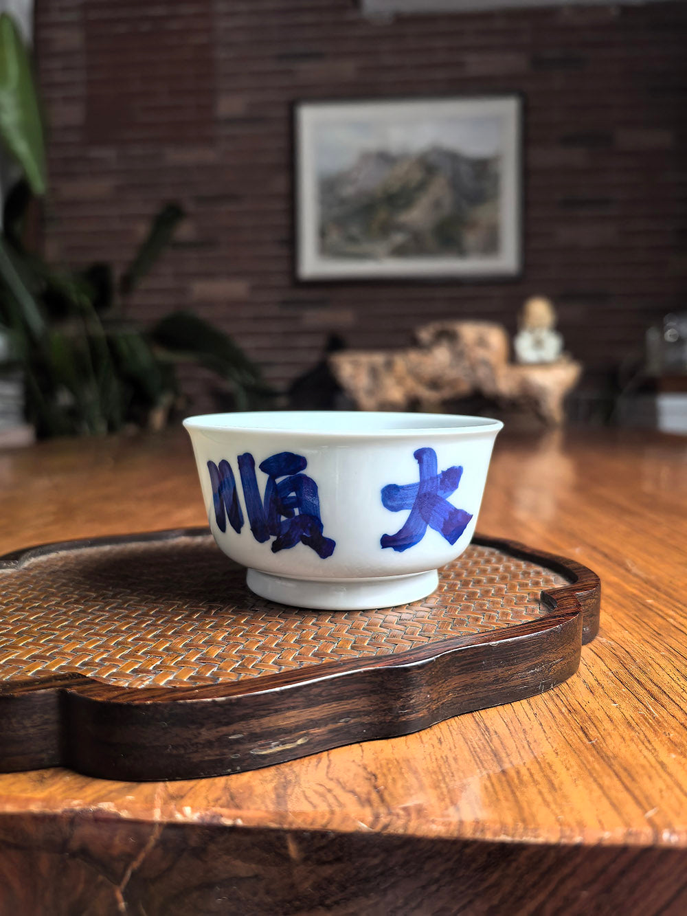 Hand-thrown Jingdezhen qingbai porcelain flat tea cup with "Great Smoothness" calligraphy, placed on a dark wood tea table with bamboo whisk background