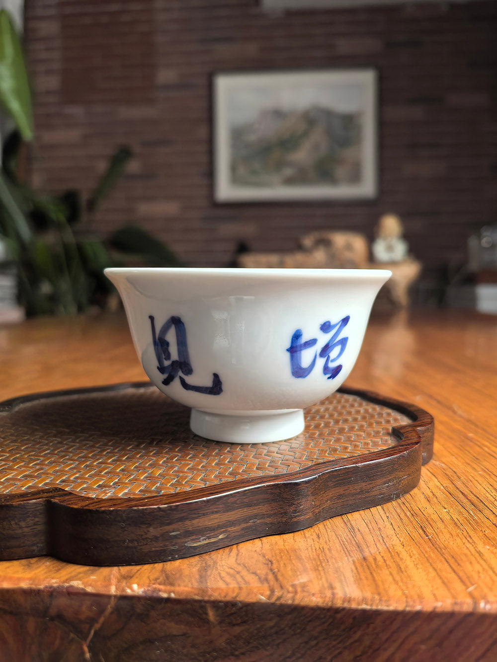 Hand-thrown Jingdezhen qingbai porcelain press-hand tea cup with "Broad Learning Expands Insight" calligraphy, placed on a dark wood tea table with bamboo mat background